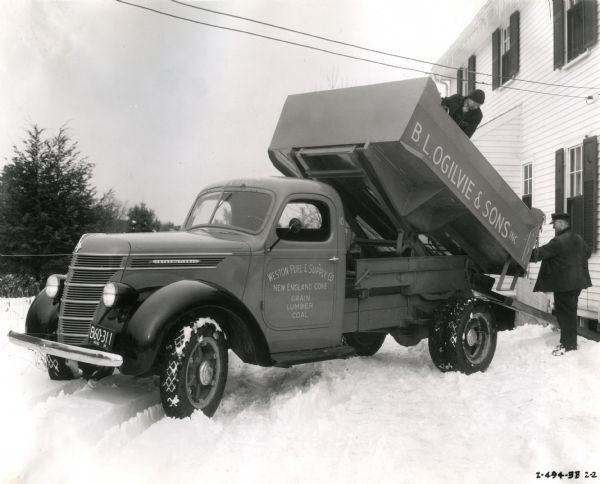 Two men use an International coal dump truck owned by B.L. Ogilvie & Sons Incorporated to transfer fuel from the bed of the truck down a chute to a residential basement. The text on the side of the truck reads: "Weston Fuel & Supply Co. New England Coke. Grain. Lumber. Coal." Snow is on the ground.