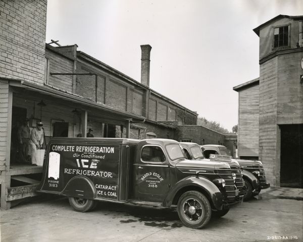 Three of Cataract Ice and Coal Company's fleet of six International D-30 trucks are parked along the company's loading dock where men stand. The text on the side of the foremost truck reads: "Complete Refrigeration Only With an Air Conditioned Ice Refrigerator. Phone 3183. Cataract Ice & Coal. Harold Esslie. Distributor. Phone 3188."