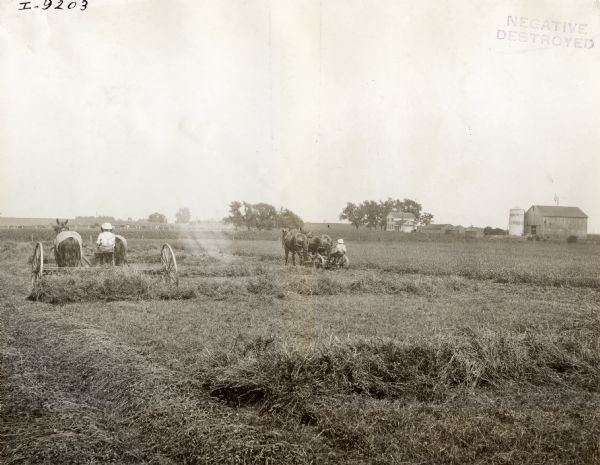 Two men are using teams of horses to pull a hay rake on the left, and  a mower on the right. There are farm buildings and cows beyond a fence in the background.