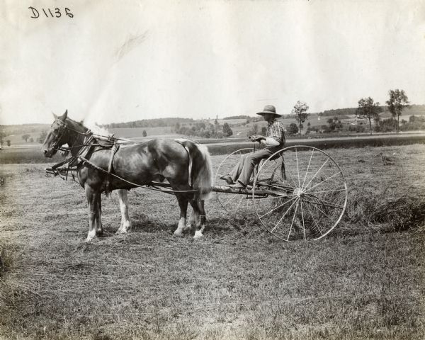 Side view of a man wearing a straw hat operating a horse-drawn hay rake in a field. In the background are hills, fields and farm buildings.