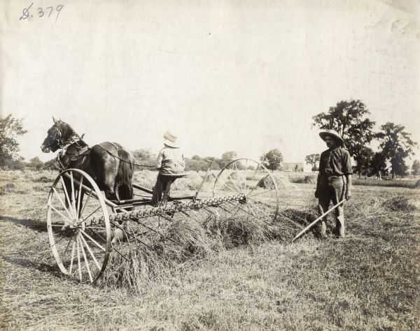 Rear view of young child operating a horse-drawn hay rake while a man is standing nearby.