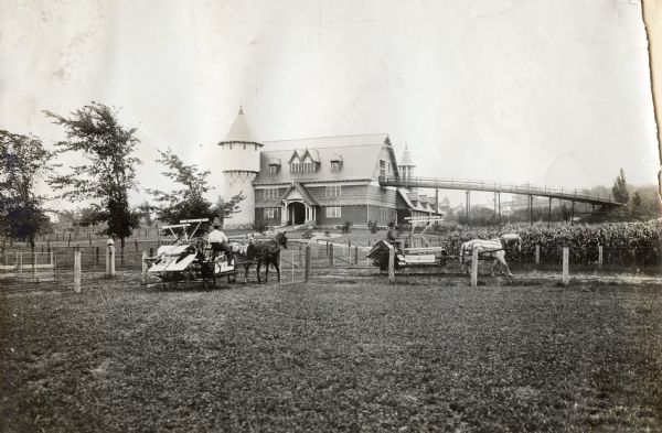Two men use horse-drawn McCormick grain binders in a field in front of a dairy barn at the University of Wisconsin-Madison. A grain elevator or walkway extends from the barn's loft.