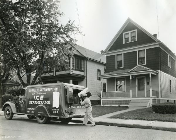 A man unloads a block of ice from the back of an International D-30 truck owned by Cataract Ice and Coal Company. The truck is parked along a curb in a residential area.