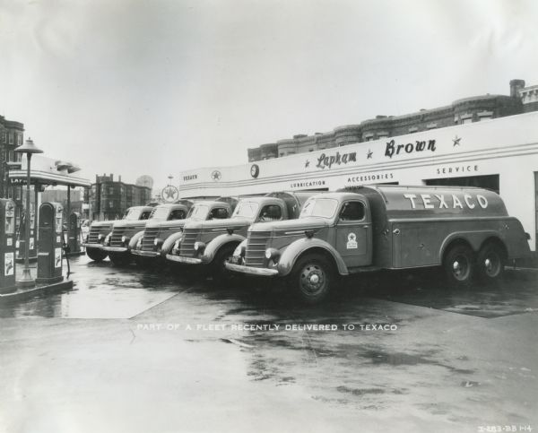 A fleet of international D-346-F trucks owned by Texaco parked outside the Lapham Brown Texaco service station. The text on the photograph reads: "Part of a Fleet Recently Delivered to Texaco." Gas pumps are on the left.