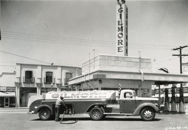 A man delivers gasoline to a Gilmore service station using an International D-40 truck. Gas pumps are in the background.