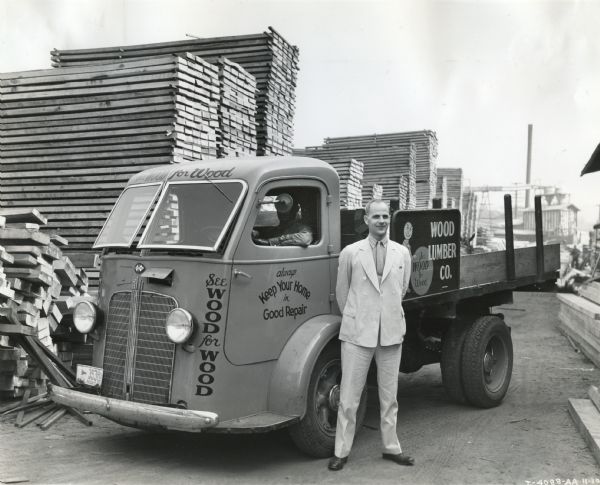 Mr. Allan K. Wood stands beside an International C-300 truck owned by Wood Lumber Company. Piles of lumber are stacked in the background, and there is a man in the driver's seat of the truck.