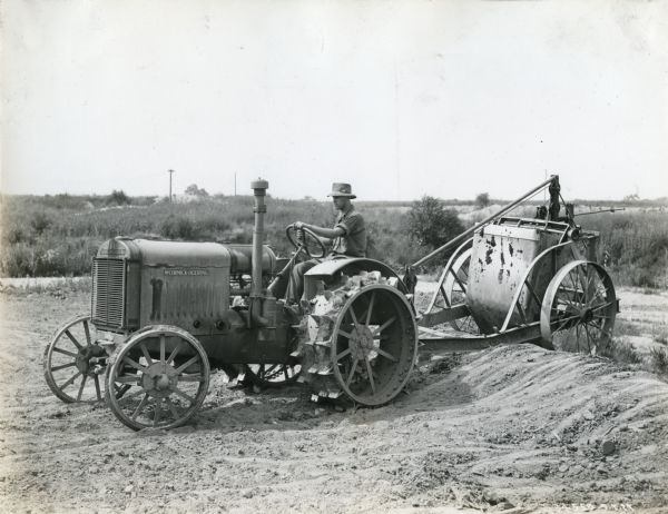A man uses a McCormick-Deering 10-20(?) tractor to pull a piece of equipment (possibly a sprayer) across a farm field.