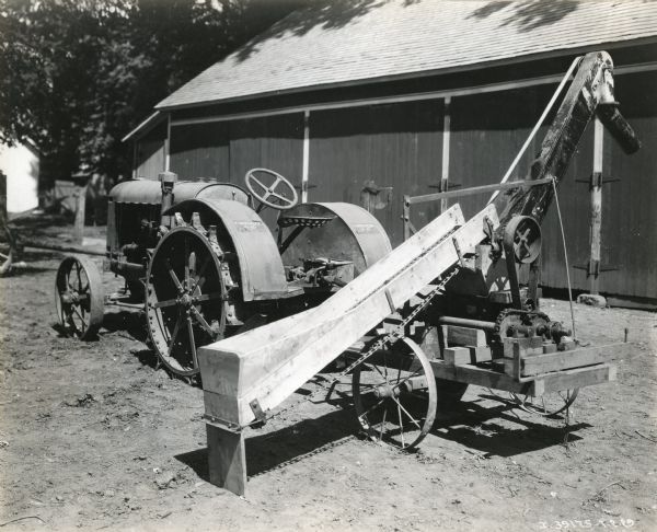 A corn husker shredder stands outside a barn in a farmyard beside a McCormick-Deering 10-20 tractor.