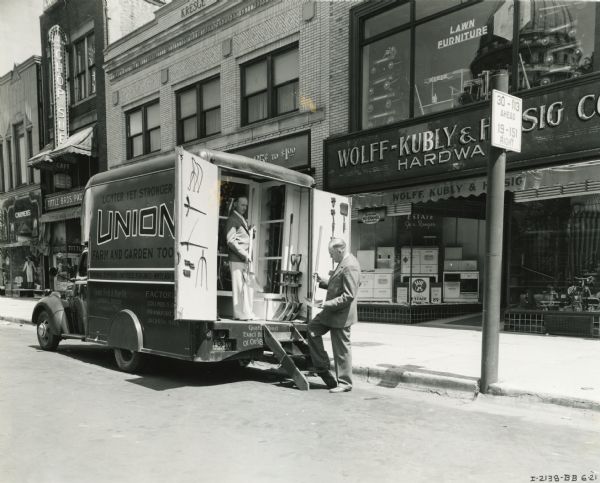 Two men look at shovels stored in the back of an International D-15 truck owned by Union Farm and Garden Tool Company. The truck is parked along the curb in front of the Wolff-Kubly & Hersig Company hardware store, located at 17 South Pinckney Street. The Wisconsin State Capitol building is seen as a reflection in a second story window.