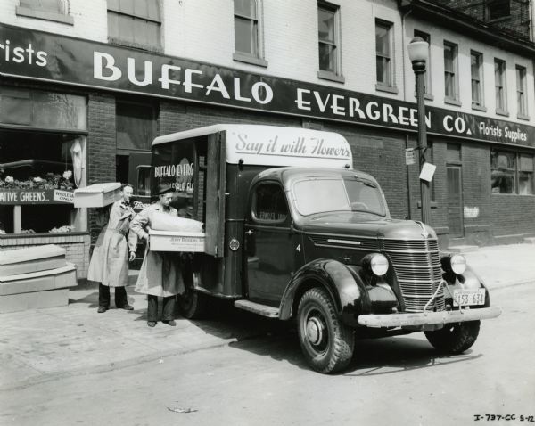 Two men load boxes into the side of an International D-15 truck owned by the Buffalo Evergreen Company, seller of florist supplies. The truck is parked outside the storefront.