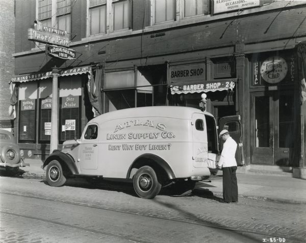 A man removes a package from the back of an International D-15 truck owned by Atlas Linen Supply Company. The vehicle is parked along the curb in front of several commercial buildings including the Konigsberg Inn, a barber shop, and the Imperial Hall Finnish Workers Club.