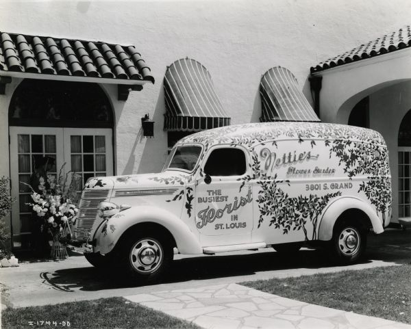 An International D-15 truck owned by Nettie's Flower Garden parked in front of a building with a tiled roof entrance and awnings over the windows. There is a delivery man standing in front of the double doors with a large flower display.