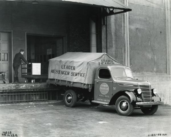 An International D-15 truck owned by Leader Messenger Service is backed up to a loading dock, and a man on the dock is pushing a wheeled cart.