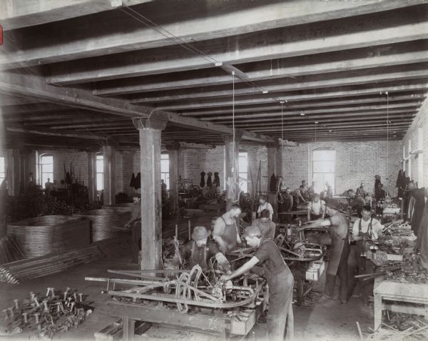 Elevated view of men working in the metal finishing room of the McCormick Reaper Works factory. Metal parts are stacked on the floor and the room is supported by heavy beams. Tall windows line brick walls.