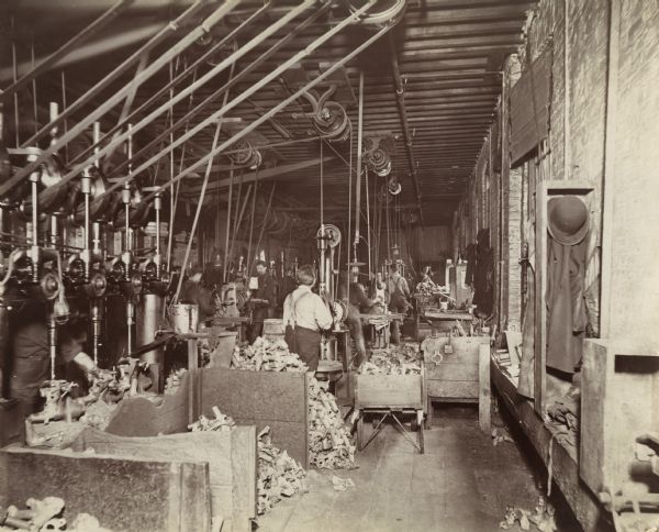 Factory workers producing farm implement parts at the McCormick Reaper Works. A pulley system is set up on the ceiling and several piles of metal pieces are separated into bins on the floor. A worker's coat and hat are hanging in the right foreground.