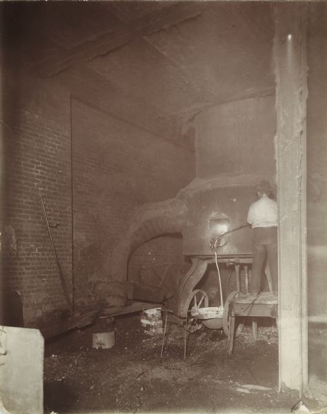 A factory worker, facing away from the camera, is standing on a wooden bench and is using a tool to reach into a furnace at the McCormick Reaper Works. Molten metal is streaming out into a crucible below.