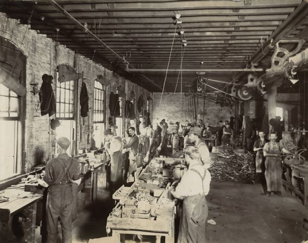 "Knotter men" work at tables in the McCormick Twine Mill. Windows line one wall of the brick room, and a system of belt-driven machinery is arranged along the ceiling.
