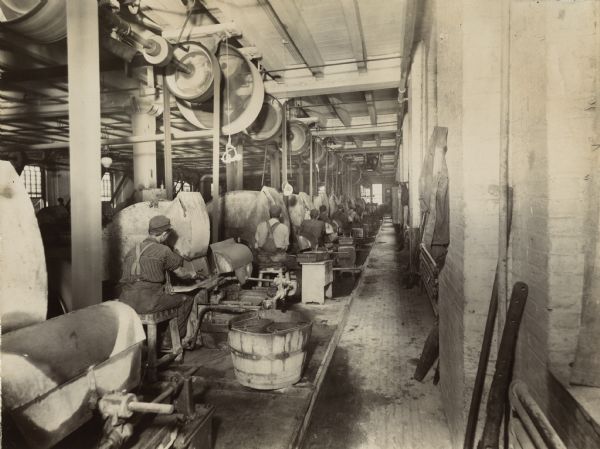 View down an aisle of male factory workers seated at machines in the McCormick Reaper Works. The machines are belt-driven from the ceiling.