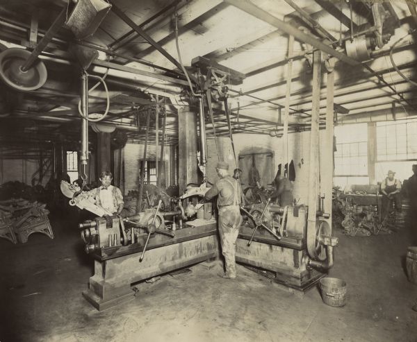 Factory workers use a belt-driven universal boring machine at the McCormick Reaper Works. Additional workers stand near a wall lined with windows in the background.