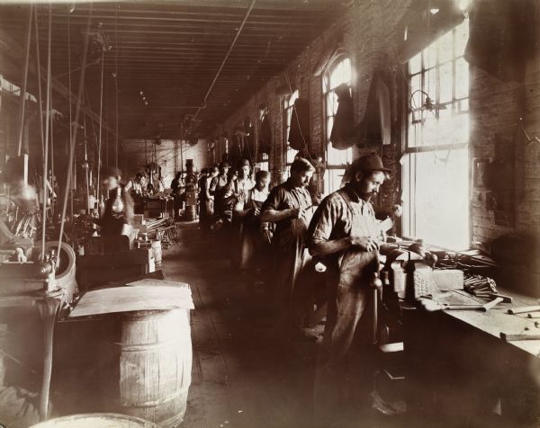 Male workers standing at stations positioned along a wall of windows inside the McCormick Reaper Works. A belt-driven power system is suspended from the ceiling at left.