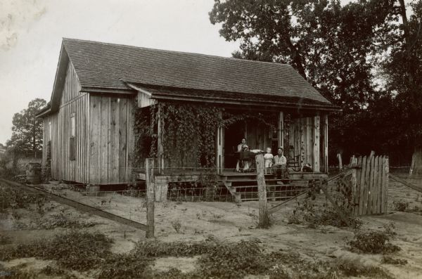 Exterior view of a wooden house, with a family, including a man, woman, dog and child on the porch. The woman is using a sewing machine near the open front door.