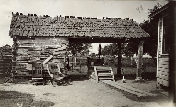 Farm structure with wood boards askew on the outer walls, and loose shingles on the roof. Underneath an open section is a hand-pump on a platform. Ducks are loose in the yard.