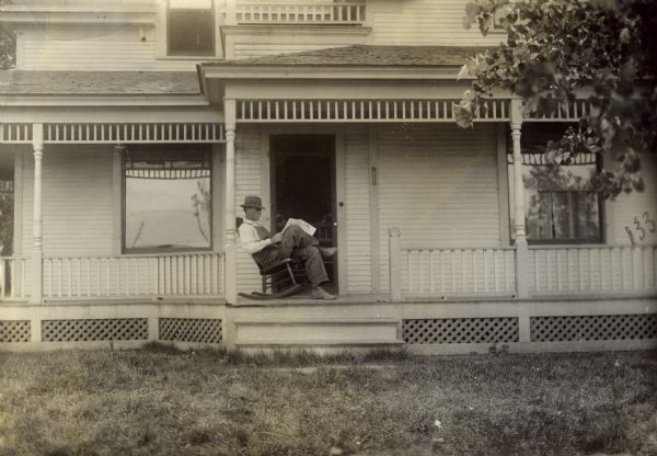 View from yard of man sitting and reading a newspaper or magazine in a rocking chair on the porch of a house.