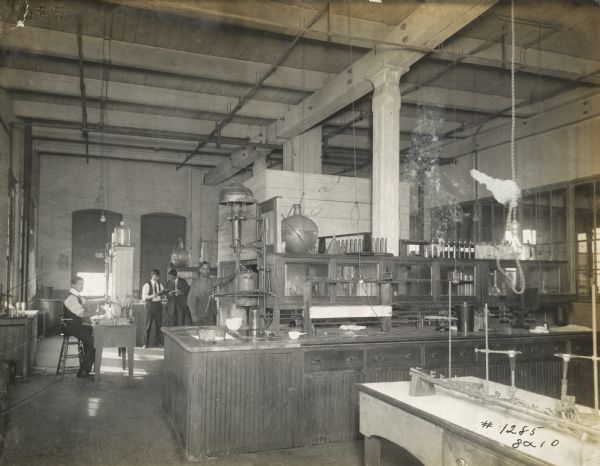 Technicians standing and sitting in the paint(?) laboratory at the McCormick Reaper Works factory. Tables are equipped with burners, beakers and other supplies.