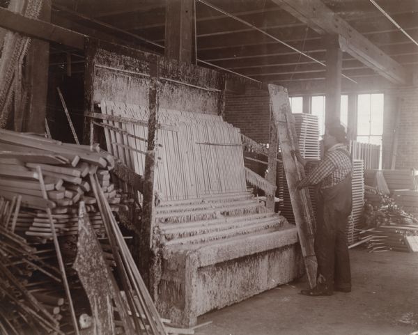 A factory worker stands beside a piece of machinery loaded with wooden pieces at the McCormick Works. Piles of wooden stakes are stacked in the foreground and background and the man holds holds what appears to be a lid by a handle.