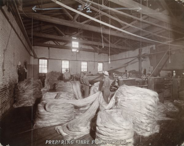 Factory workers stand with piles of sisal fiber used in the production of binder twine. The fiber is fed into spreading machines on the right.
