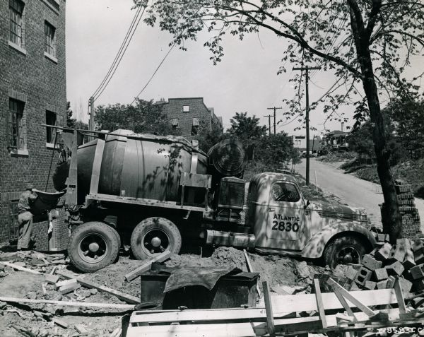 An International D-246-F truck outfitted with a Jaeger 3 1/2 cubic yard concrete mixer parks at a construction site. Boards and cinder blocks are piled in the foreground and a male construction worker is standing near the rear of the truck.