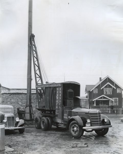 An International D-246-F truck outfitted with a Model 200 power unit operates a crane. The truck was owned by Merchants Cartage Company, Ltd. A house is in the background.