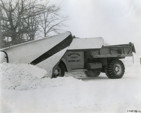 Winter scene with an International D-70 truck owned by the Waukesha County Highway Department and outfitted with a scraper clearing snow from a rural road.