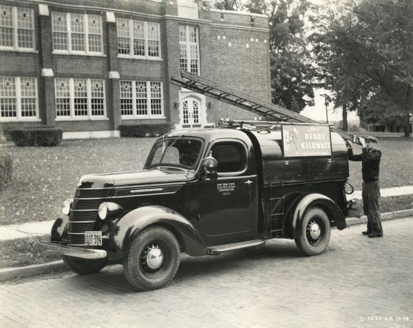 A man unloads a ladder from the top of an International D-2 truck owned by New York State Electric and Gas Corporation and used for maintenance purposes. The sign on the side of the truck reads: "Meet Reddy Kilowatt, Your Electrical Servant."