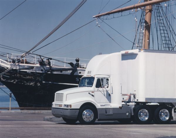 Color photograph of an International 8300 semi-truck (tractor-trailer) parked in a dock area near a sailing ship.