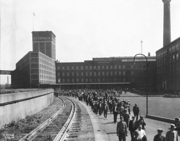 Stream of factory workers leaving International Harvester's McCormick Works. A sign along the front of the factory reads: "'Quality is the Foundation of our Business'! Alex. Legge, President."