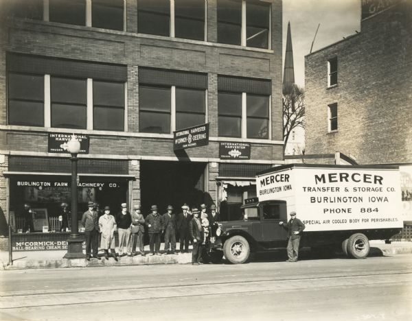 A group of men stand outside the Burlington Farm Machinery Company storefront where an International H-5 truck owned by the Mercer Transfer Company is parked by the curb. The truck featured a 190-inch wheelbase and an air-conditioned body. The Burlington Farm Machinery Company was an International Harvester dealership.