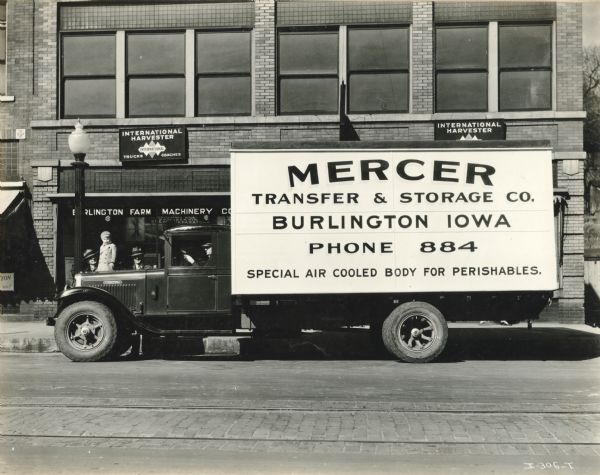An International H-5 truck with a 190" wheelbase and a refrigerated body parked in front of the Burlington Farm Machinery Company, an International Harvester dealership. The truck was owned by the Mercer Transfer & Storage Company. There is a driver in the driver's seat, and two men with a young child stand on the sidewalk behind the truck.