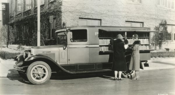 Two women select books from the opened back of a traveling branch library in a truck parked on a street.
