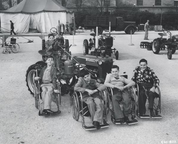 Four boys in wheelchairs pose in front of a McCormick-Deering Farmall C. Another boy sits behind the steering wheel. Other agricultural equipment is arranged, seemingly on display, near a tent in the background.