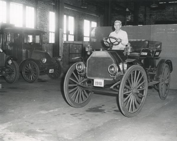 A man is sitting at the wheel of an International Auto Wagon with seats in the back. The vehicle is on the floor of an factory building, possibly the McCormick Works in Chicago. Other vintage International trucks are parked on the left.