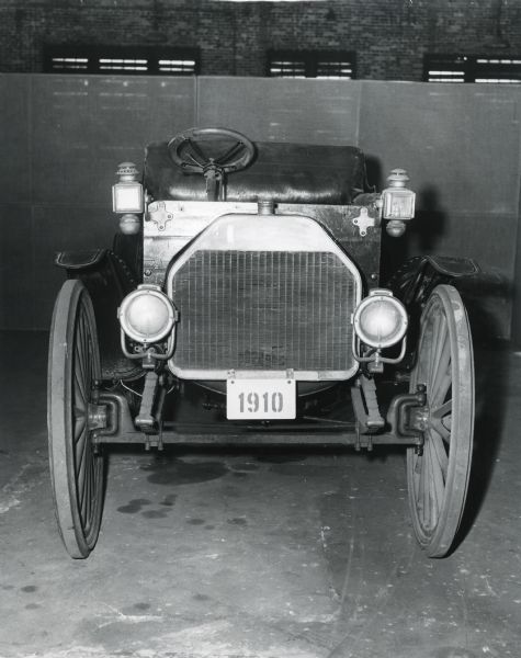 View of the front grill of an International Auto Wagon with seats in the back, parked on the floor of a factory building, possibly McCormick Works in Chicago.