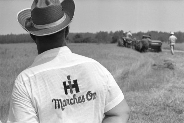 View over shoulder of a man facing away and wearing a straw hat and a shirt reading "IH Marches On" as he looks across a field on the International Harvester company farm at Tifton. In the background men are using agricultural equipment. International Harvester offered classes on the farm in order to provide its sales force with hands-on experience with agricultural equipment in the field.