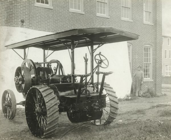 Three-quarter rear view of an Otto gasoline engine tractor, taken in front of a white backdrop held on one side by a man standing at right in the grass in front of a brick building.