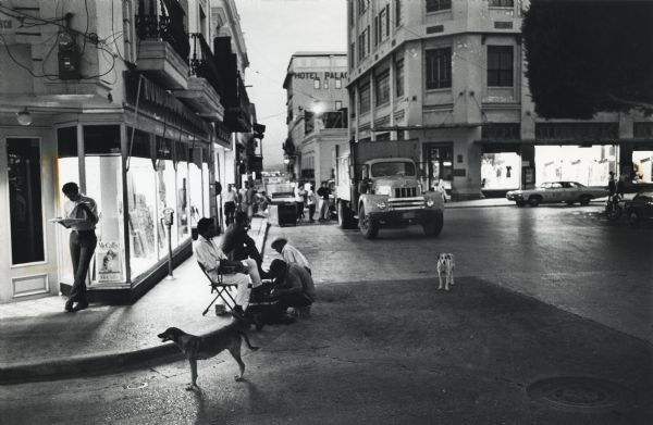 City street in Puerto Rico, seemingly at dusk. Shop windows are brightly lit, and a man leans against one while reading a newspaper. Other men are having their shoes shined on a sidewalk, and two dogs are standing in the street nearby. In the background, an International garbage truck is parked in the street.