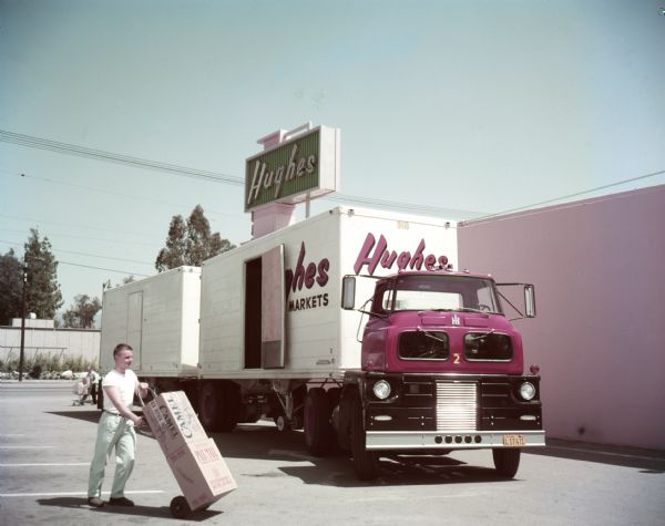 A man pushes boxes of Pall Mall and Camel cigarettes on a handcart in front of an International ACO Sightliner truck with an open side door. The ruck was owned by Hughes Markets.