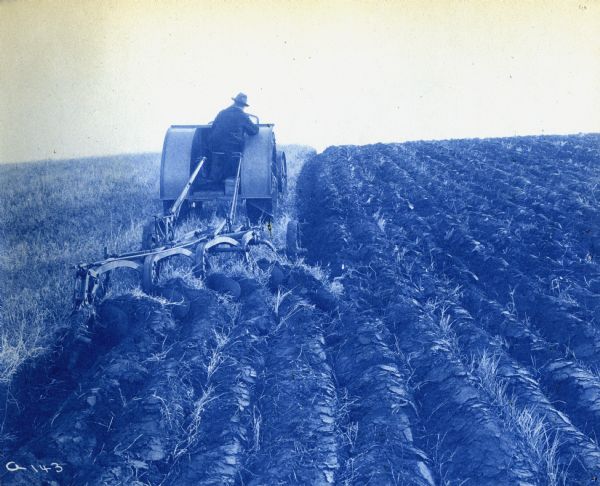 Cyanotype print of a farmer using an Andrews-Kinkade Model D four-plow tractor to work in a field.