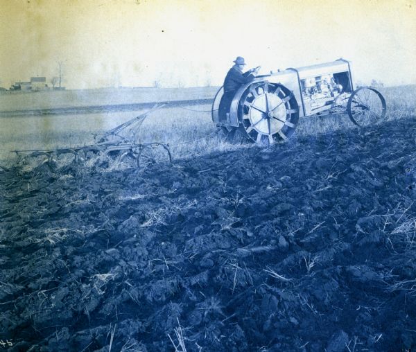 Cyanotype print of a man using an Andrews-Kinkade Model D four-plow tractor to work in a field. Farm buildings are in the background at left.