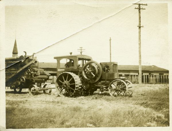 A man sits behind the wheel of an Avery tractor pulling a piece of farm machinery. In the background is a long open building with a tower. The back of the photograph reads, "Ellis. Avery's 'Infant.' Modern Power Company."