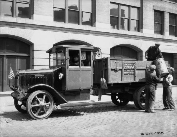View across cobblestone street towards men loading what appears to be an International garbage truck on a city street. Two men are lifting a can up towards another man standing in the bed of the truck. A fourth man is sitting behind the wheel. An American flag is attached to the front of the truck, and a sign on the side of the truck reads: "James Duffy, Jr., 555 Broome St."
